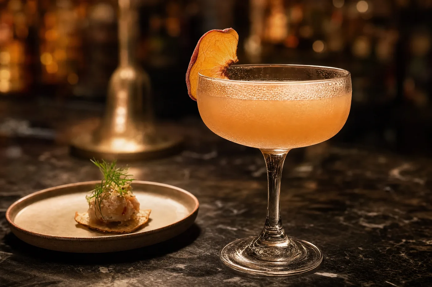 A peach cocktail in a coupe glass beside a single small bite on a plate, on a dark bar in warm light
