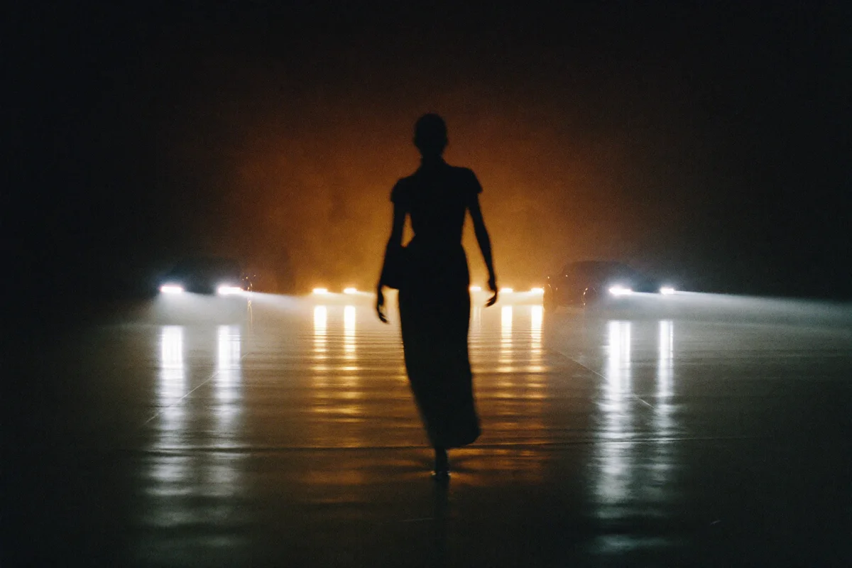 A silhouetted figure walking a runway lined with car headlights in a dark showroom