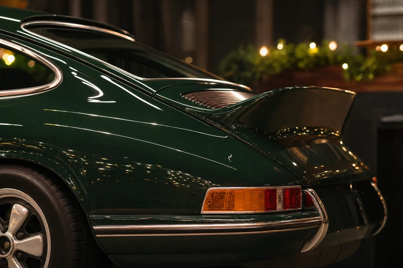 The ducktail and taillight of a dark green classic Porsche 911 with holiday garland lights softly out of focus behind it