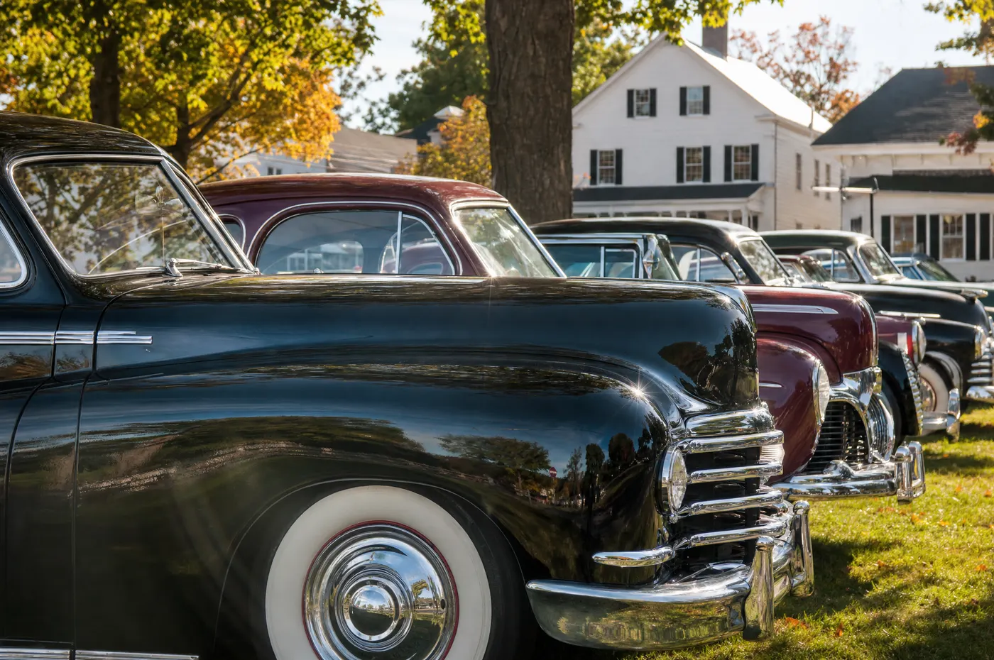A row of polished 1940s classic cars parked on a New England town green, a white colonial house behind them